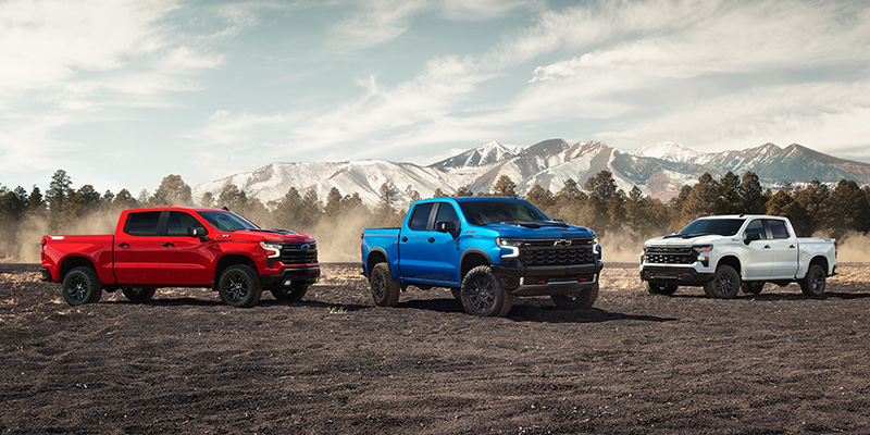 Three Chevrolet Silverado trucks in red, blue, and white parked before a mountain range