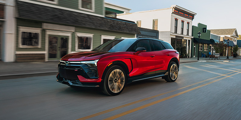 Red Chevrolet Blazer EV driving down a town main street past storefronts.